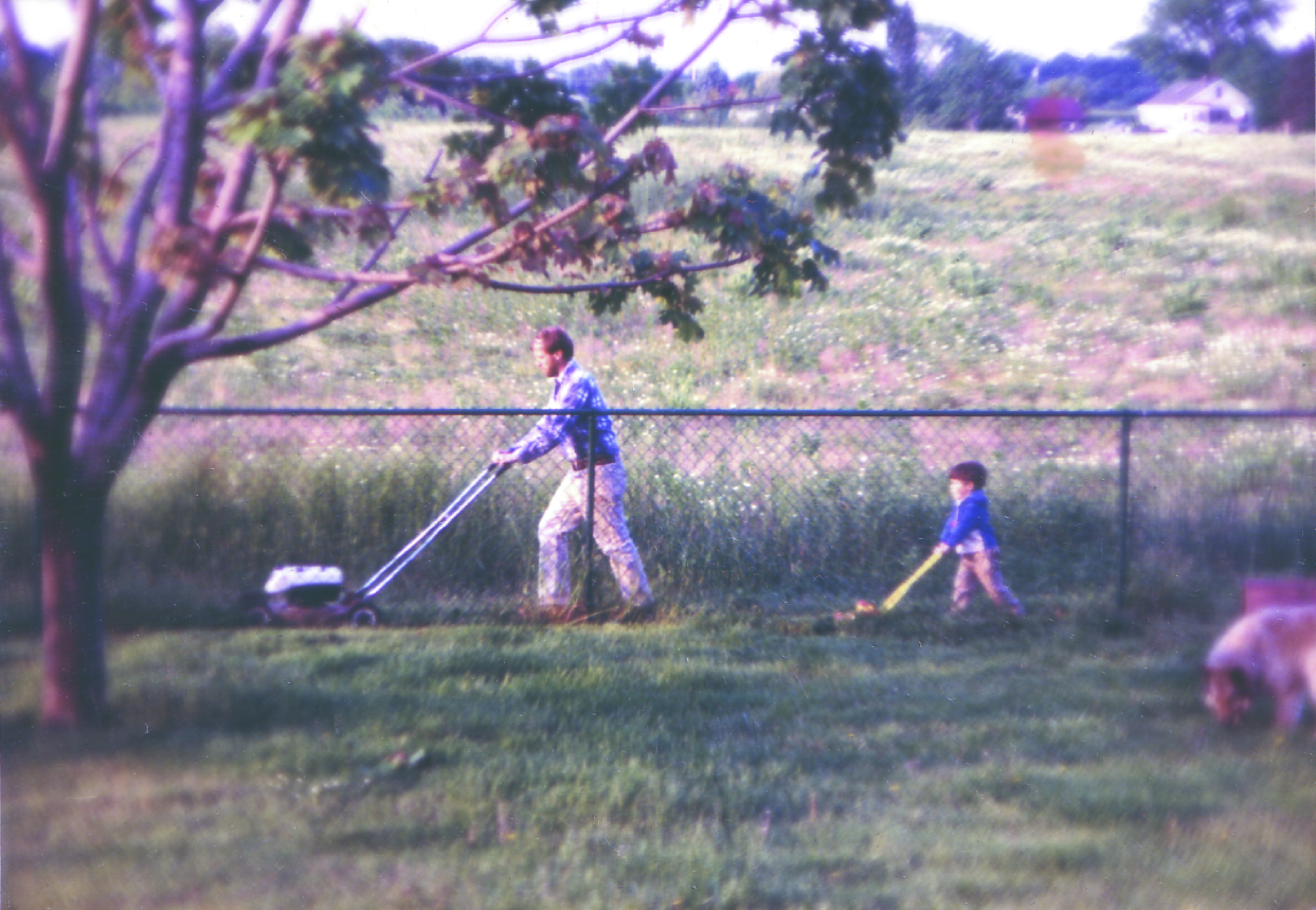 A man pushes a lawnmower with a small child behind him pushing a toy lawnmower.